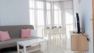 Una sala moderna con un sofá gris, una mesa de centro blanca y un comedor con mesa y sillas blancas. Un televisor está montado en un mueble de madera.