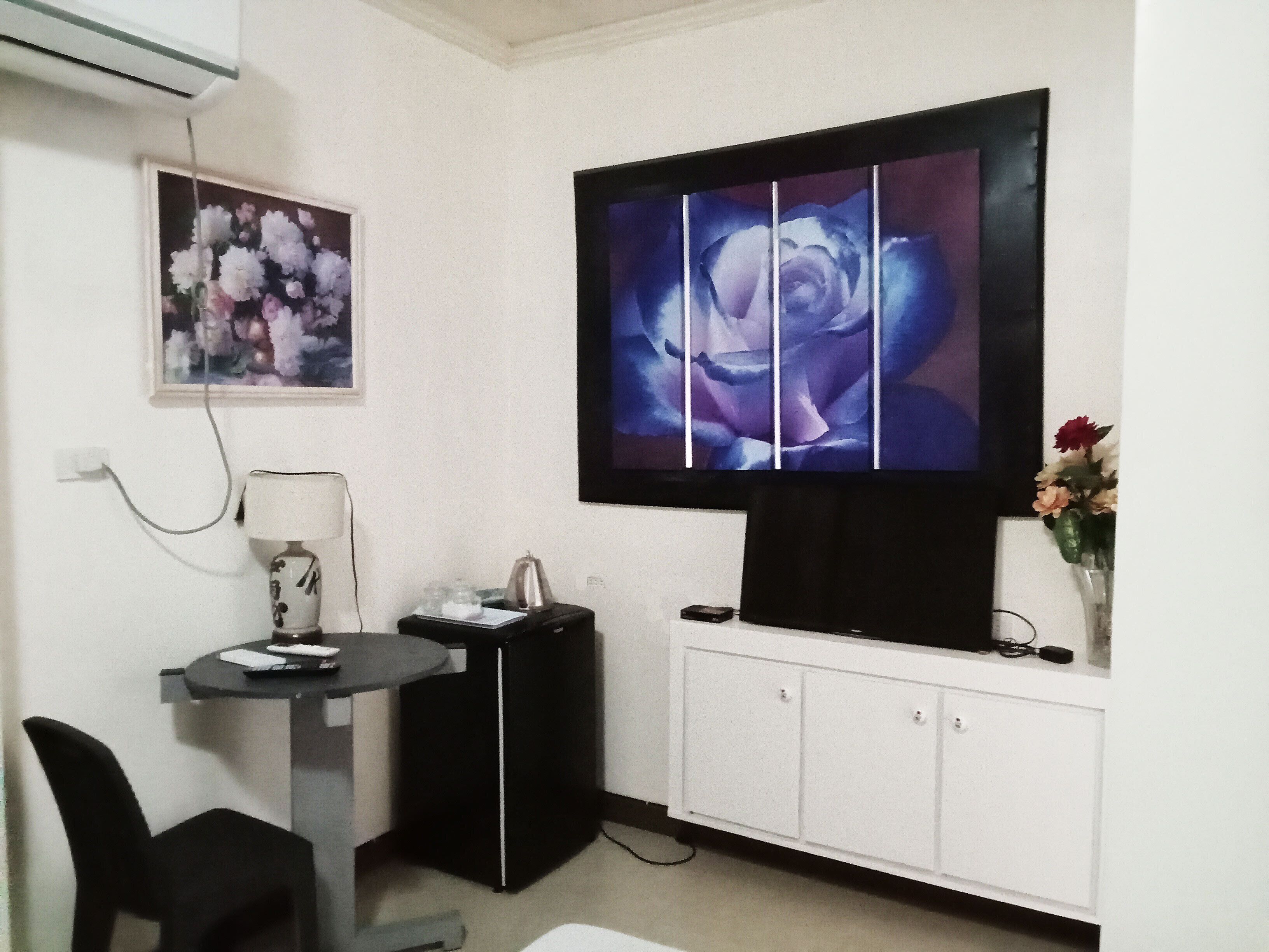 A living room with a flat-screen TV mounted on the wall, a small table with a lamp, a chair, a cabinet below the TV, and a framed floral artwork on the wall.