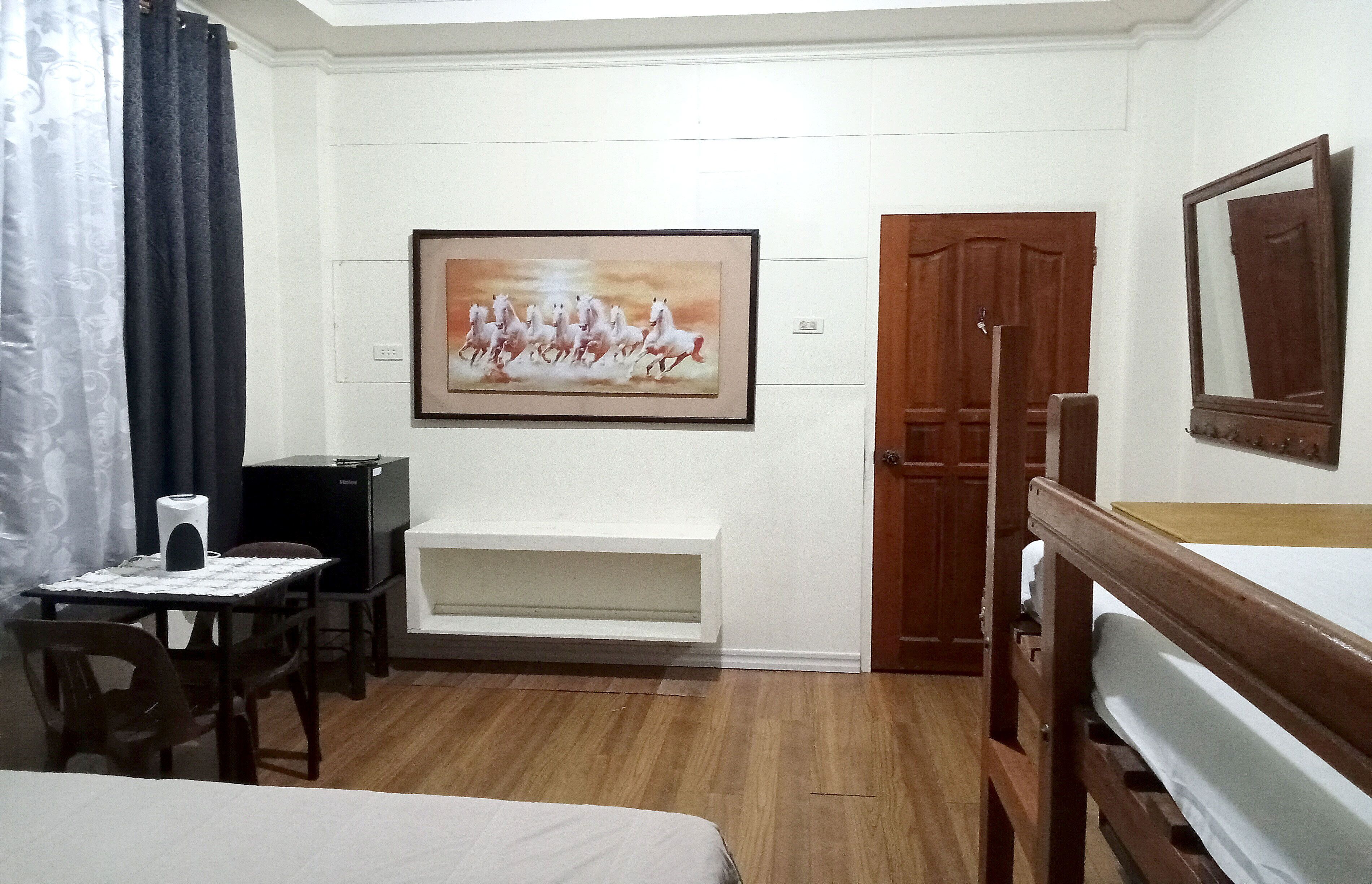A hotel room with a bed, a desk with a chair, a television, a painting, a wooden door, and a mirror.