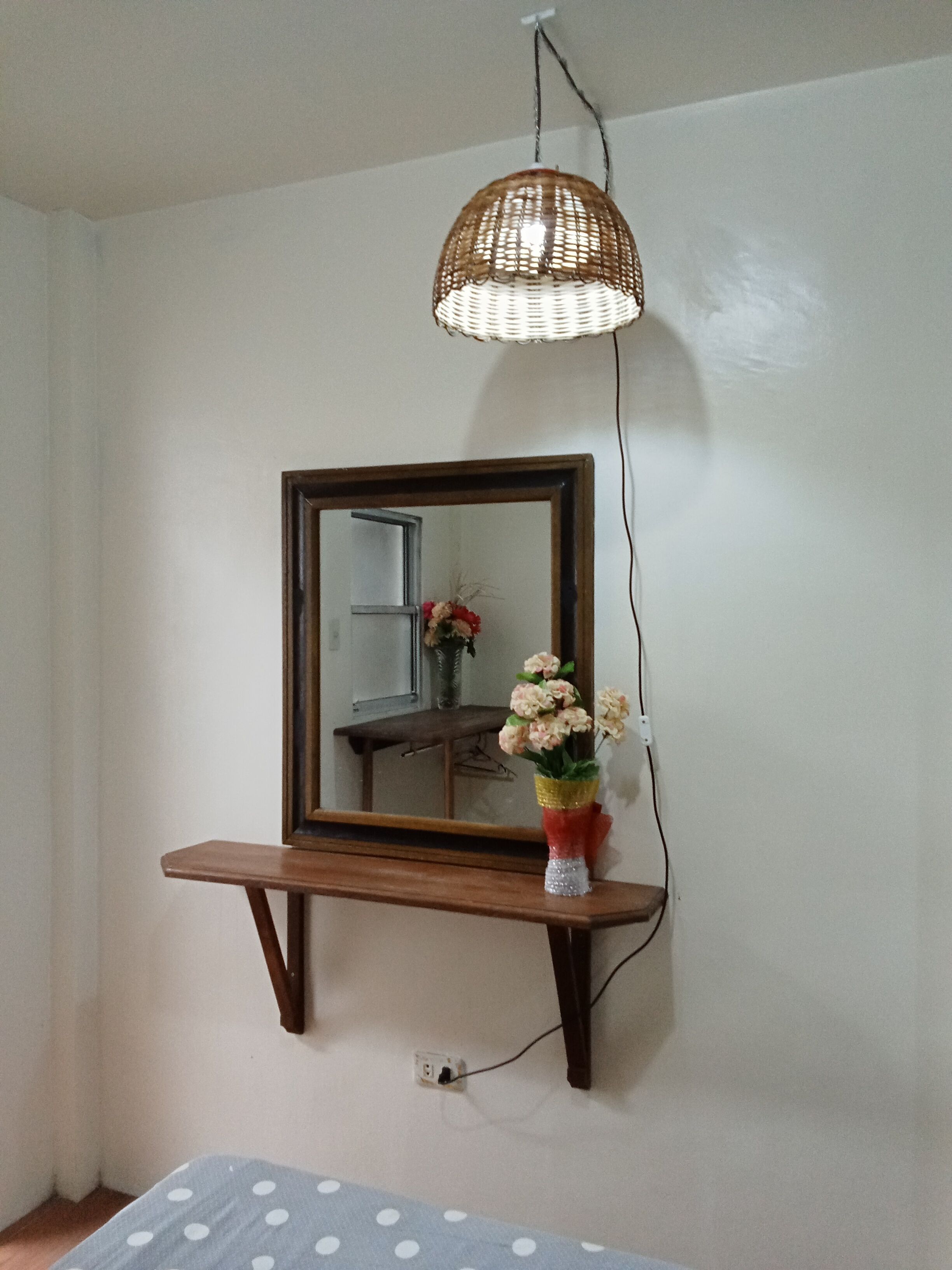 A room with a mirror on a wooden shelf, a woven light fixture, and a vase with flowers.