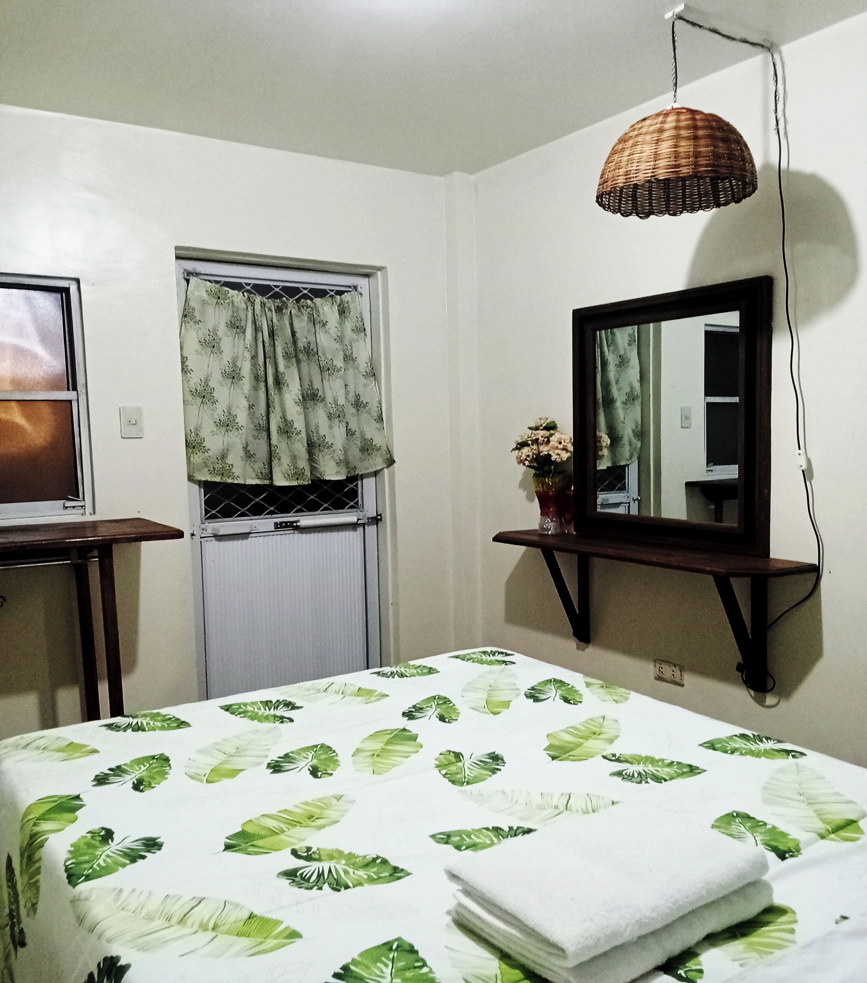 A bedroom with a bed, a window with curtains, a mirror on the wall, and a hanging light fixture.