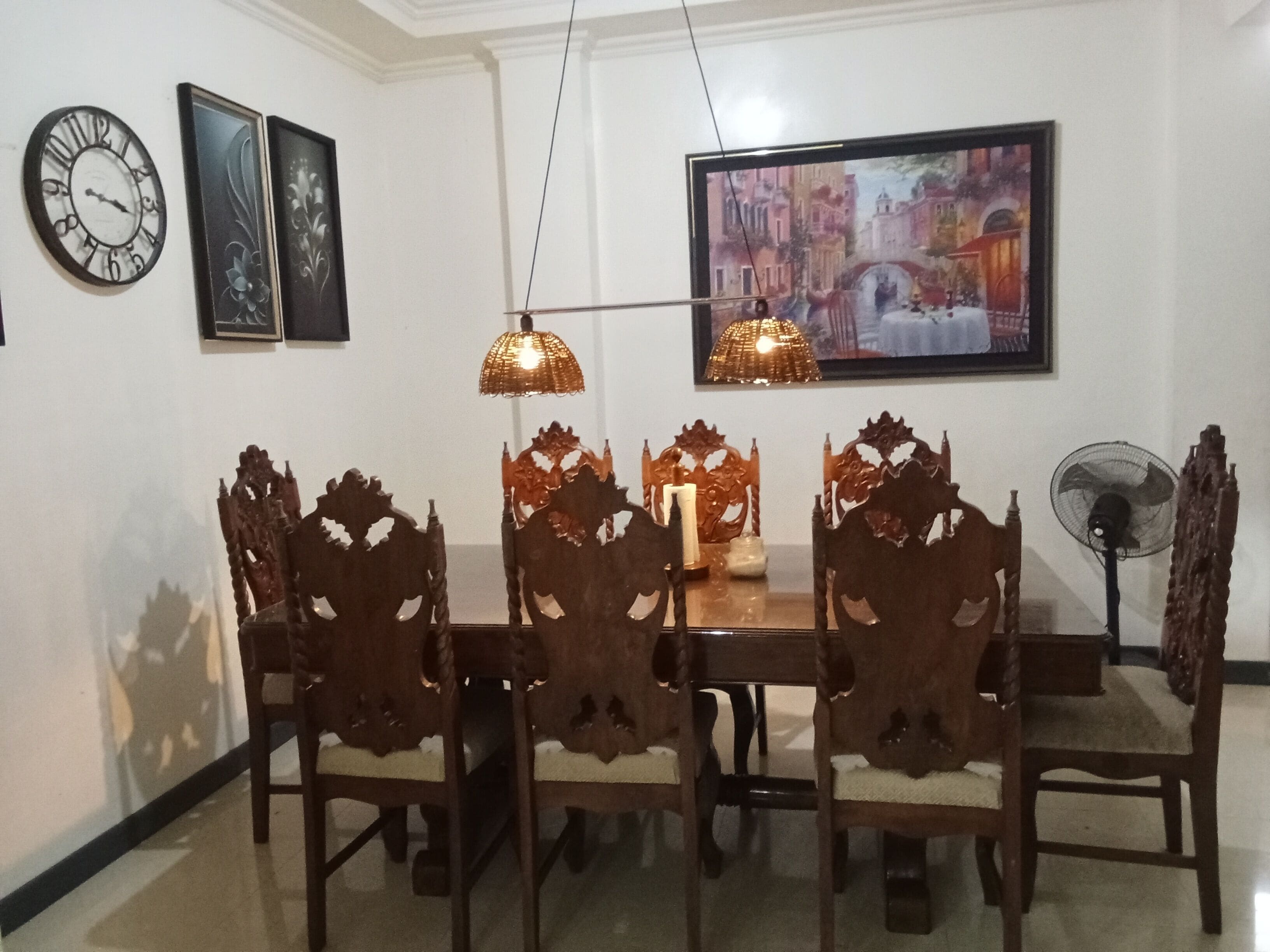 A dining area with a wooden table and chairs, a clock, framed artwork, and a painting on the wall.