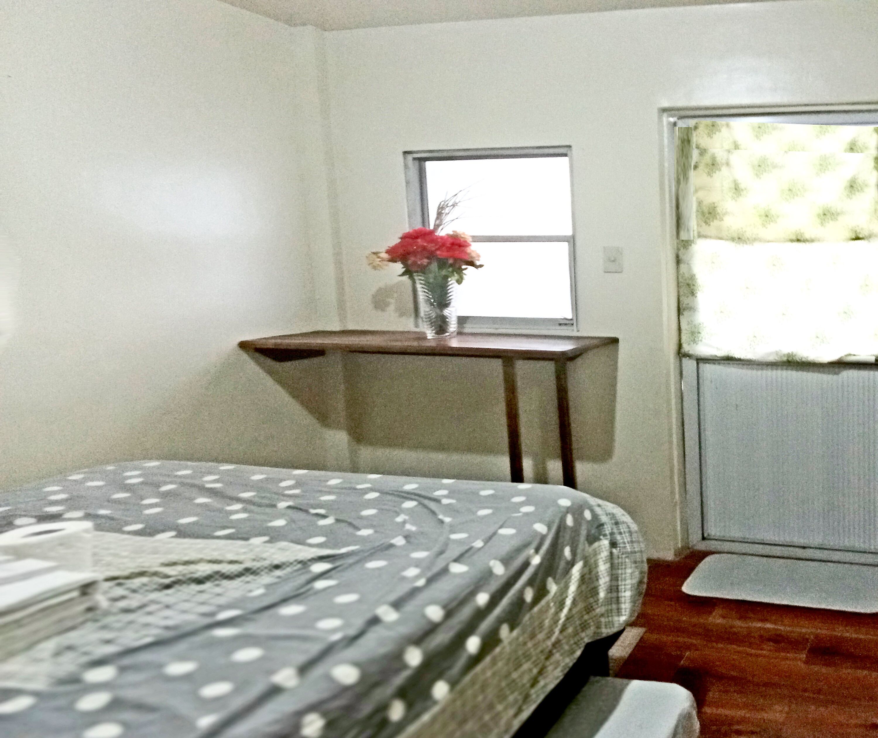 A bedroom with a bed, a small table with a vase of flowers, a window, and a door.