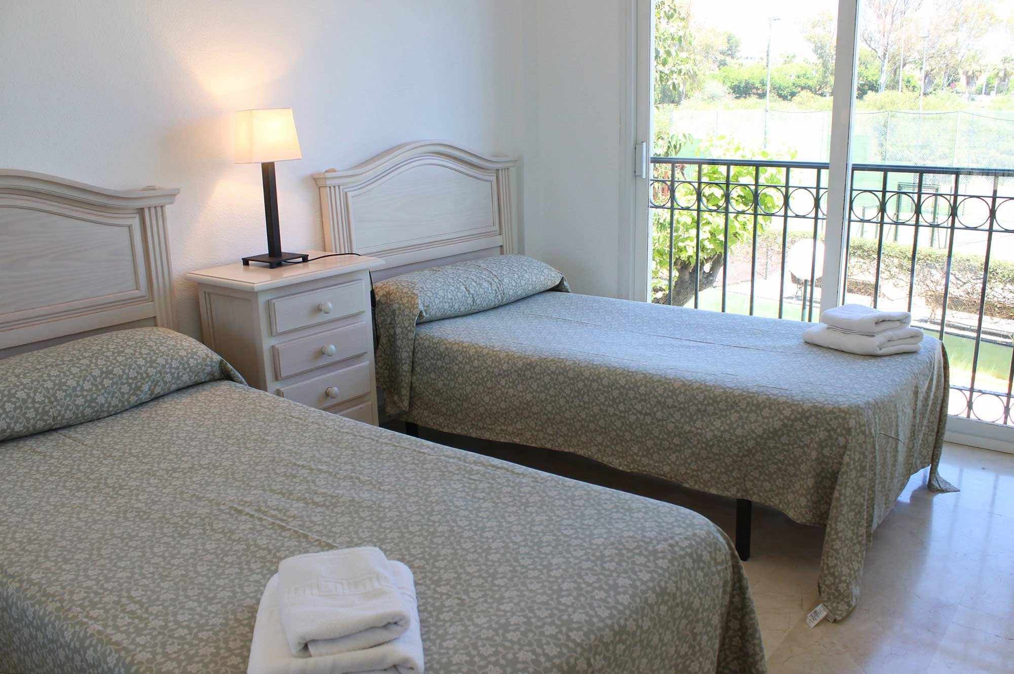 Dos camas individuales con ropa de cama floral, una lámpara de noche y vistas a un balcón con barandilla.