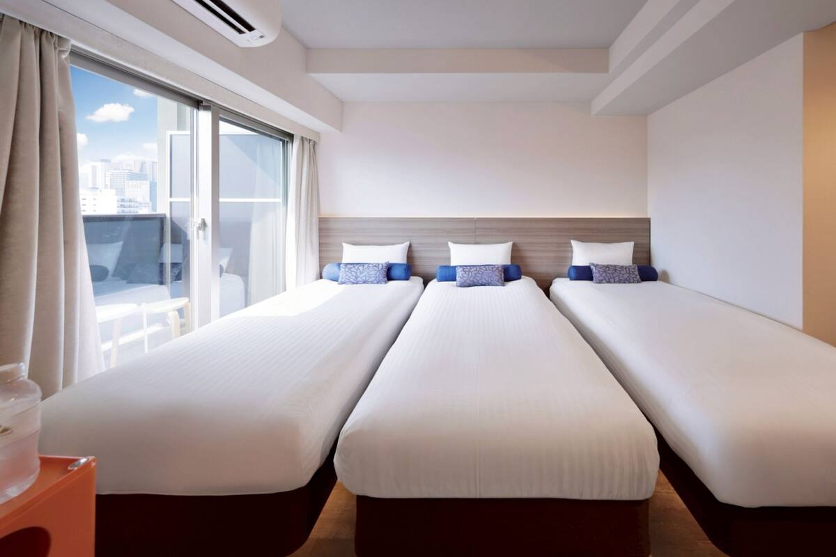 A hotel room with three single beds, a large window with curtains, and a view of the city.