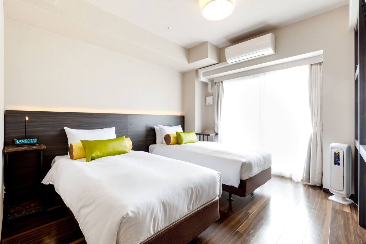 A hotel room with two beds, a nightstand with a clock, an air conditioning unit, and a window with curtains.
