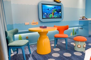 A children's playroom with a round yellow table, two chairs, a television displaying an underwater scene, and a blue carpet with white polka dots. - Dolphin Bay Hotel (Yuxi)