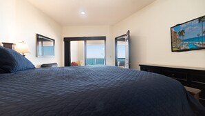 Ein Hotelzimmer mit einem großen Bett, einem Schminktisch, einem Nachttisch, einer Lampe, einem Bild von einem Strand und einer Tür zu einem Balkon mit Meerblick.