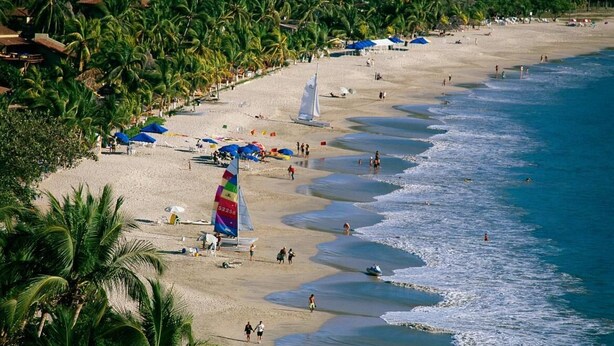 A beach with palm trees, a sailboat, and beach umbrellas. - Fabuloso Hotel Las Vegas (Zihuatanejo)
