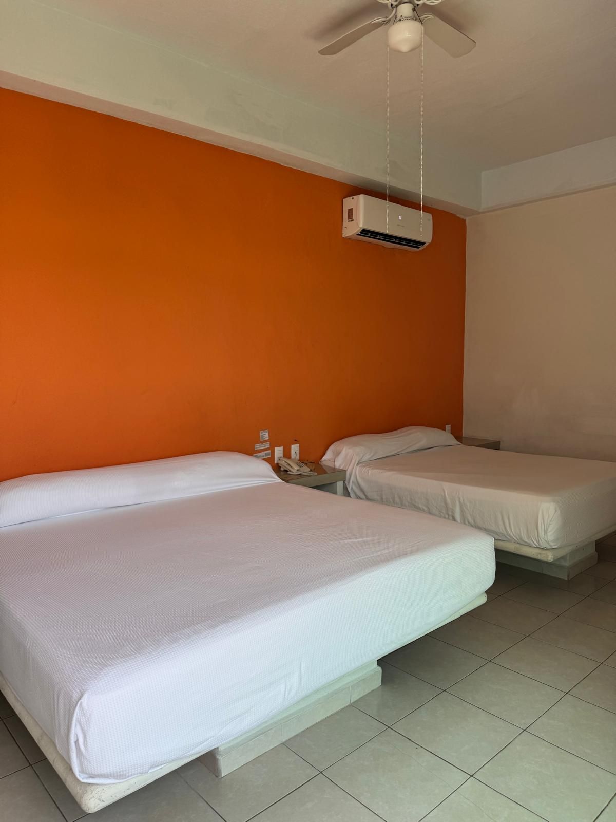 A hotel room with two beds, an air conditioning unit, a ceiling fan, and a small table with a phone.