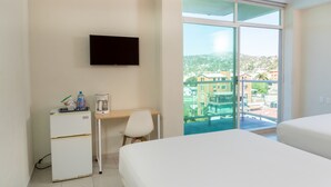 A hotel room with a bed, a desk with a chair, a TV mounted on the wall, and a large window offering a view of buildings and greenery. - Fabuloso Hotel Las Vegas (Zihuatanejo)
