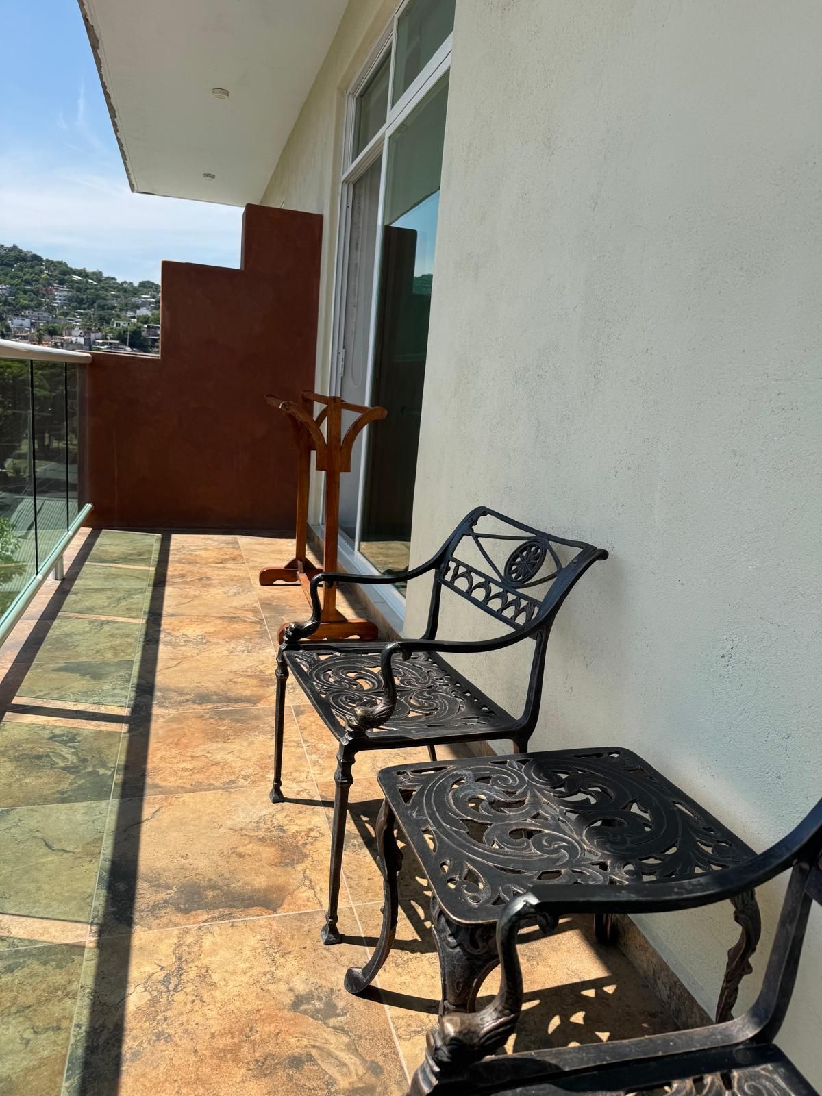 A balcony with two ornate metal chairs and a wooden chair, a glass railing, and a view of a hillside.