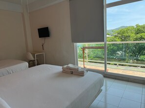 A hotel room with two beds, a TV mounted on the wall, and a large window offering a view of greenery and hills.