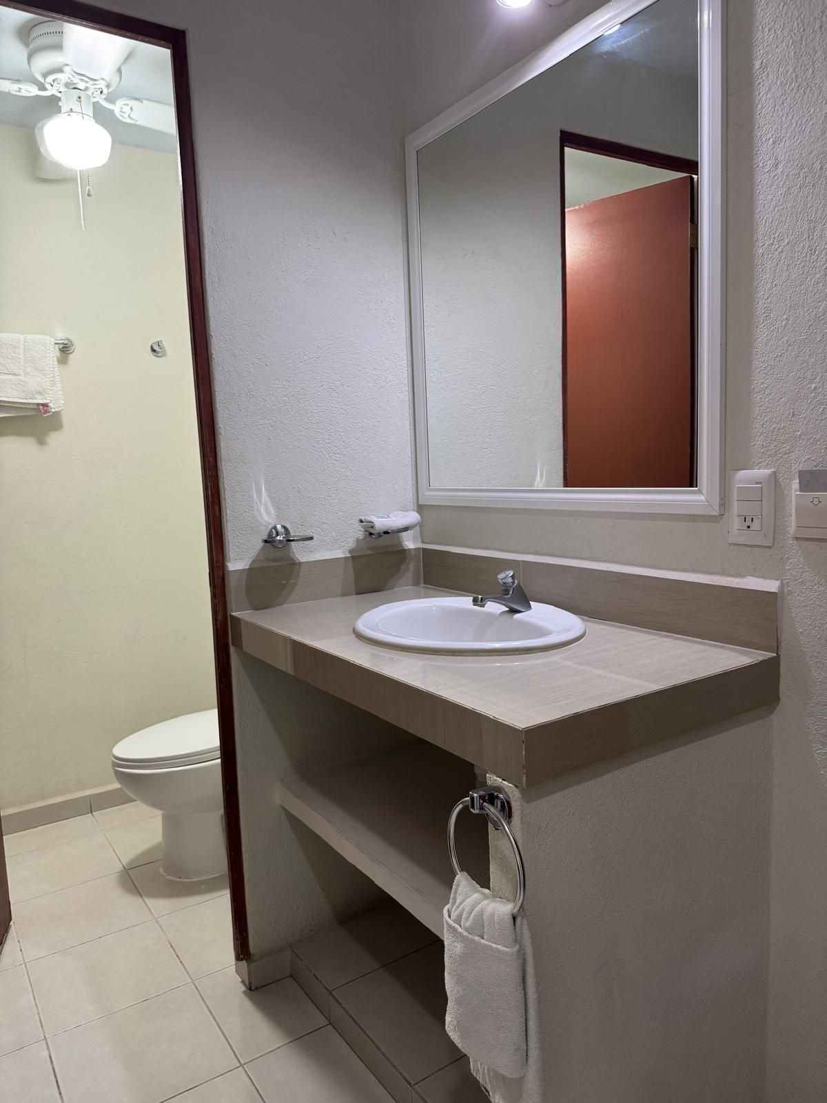 A bathroom with a white sink, a mirror above it, and a toilet visible through an open door.