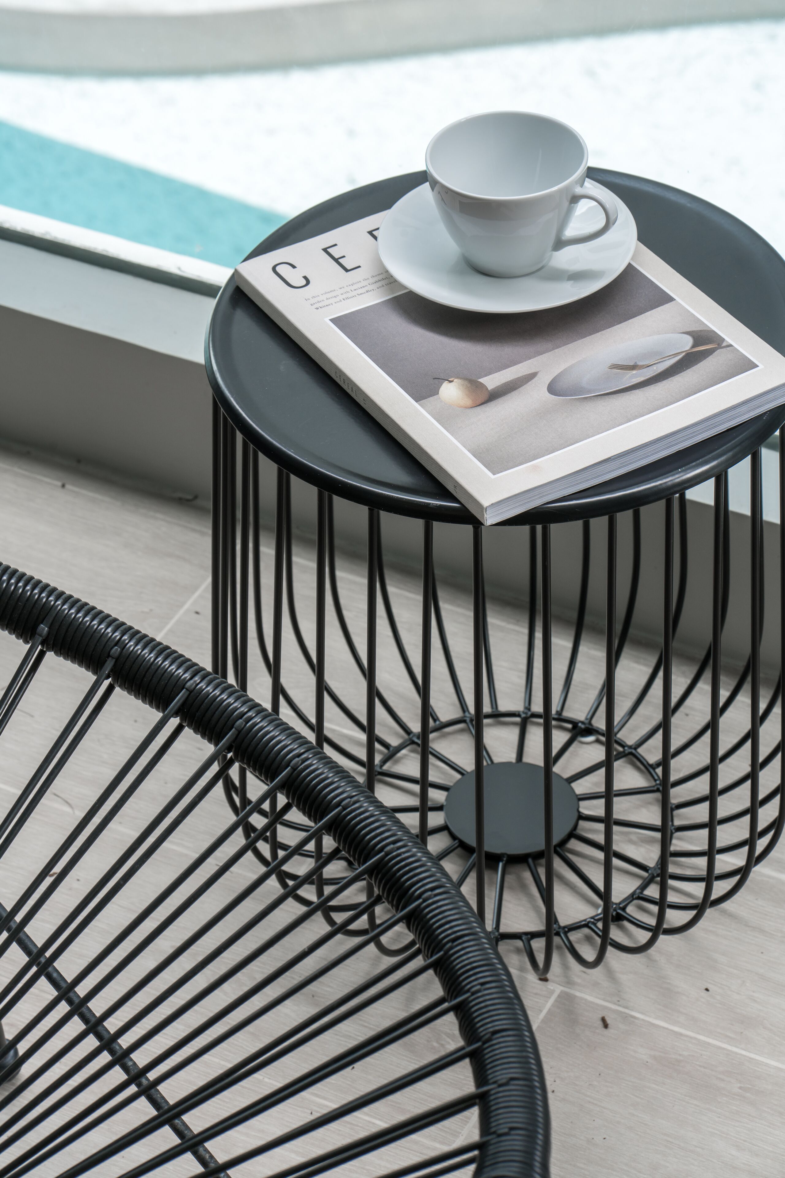 A round black metal table with a white cup and saucer on top, a magazine, and a black metal chair beside it.