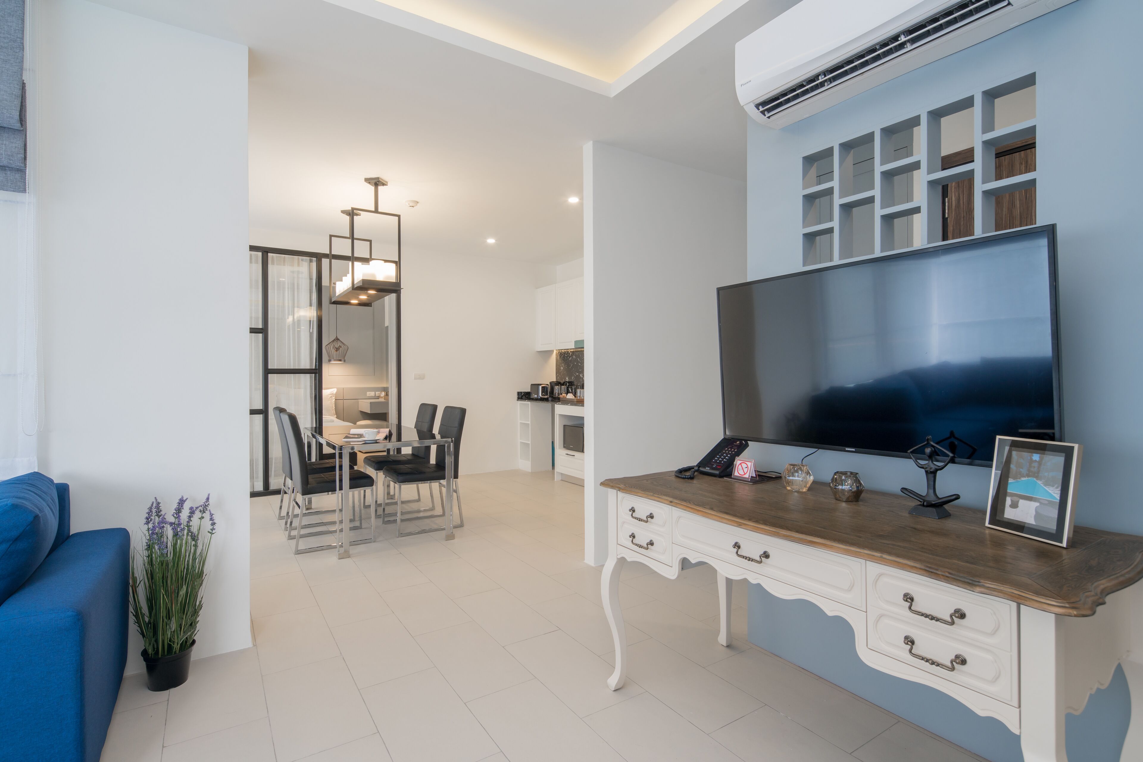 Un salon moderne doté d’un grand téléviseur à écran plat, d’un bureau en bois avec des tiroirs, d’un canapé bleu et d’un coin repas agrémenté d’un lustre.
