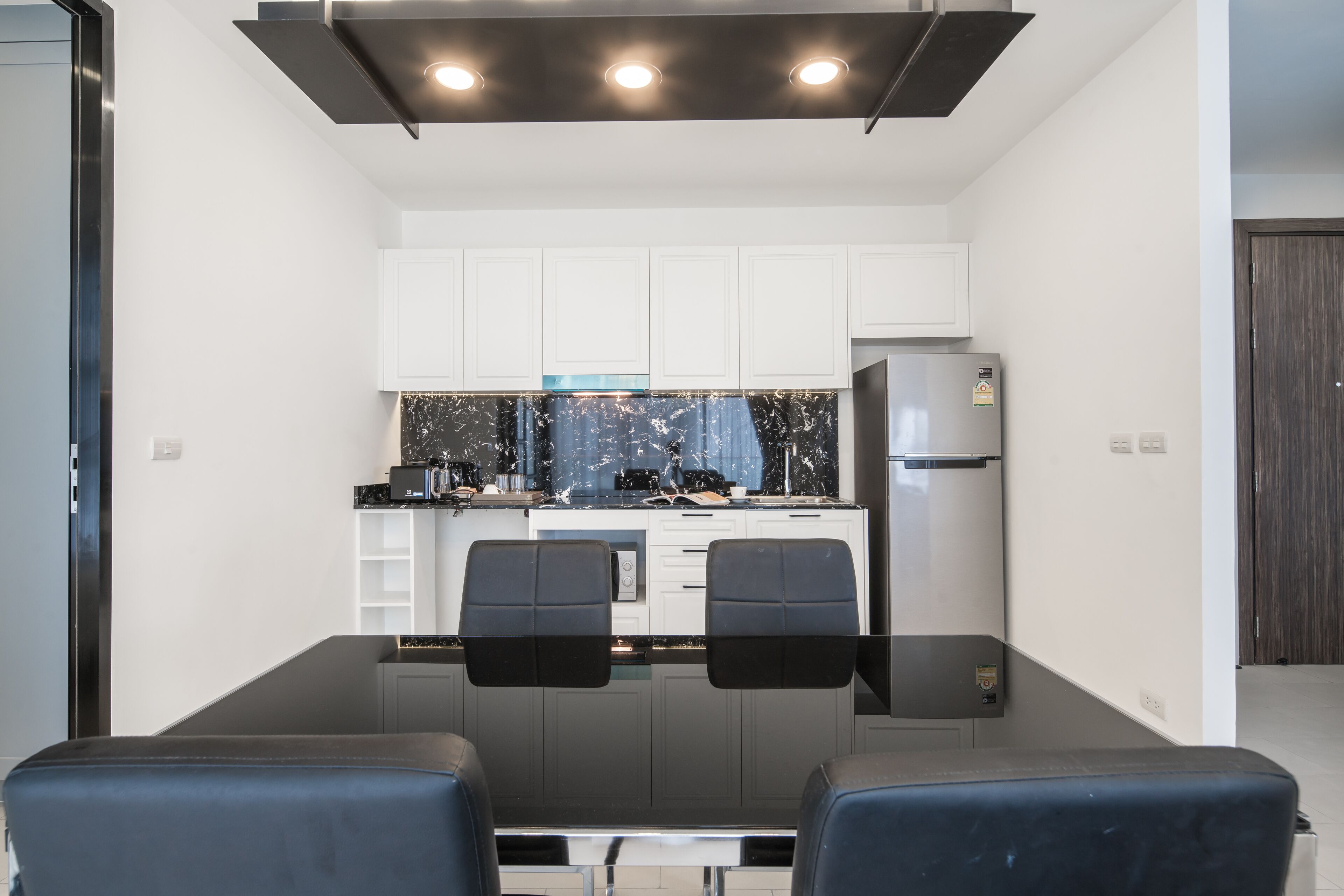 A modern kitchen with white cabinets, a black countertop, and a stainless steel refrigerator. There are two black chairs at a table in front of the kitchen.