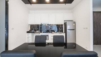 A modern kitchen with white cabinets, a black countertop, and a stainless steel refrigerator. There are two black chairs at a table in front of the kitchen.