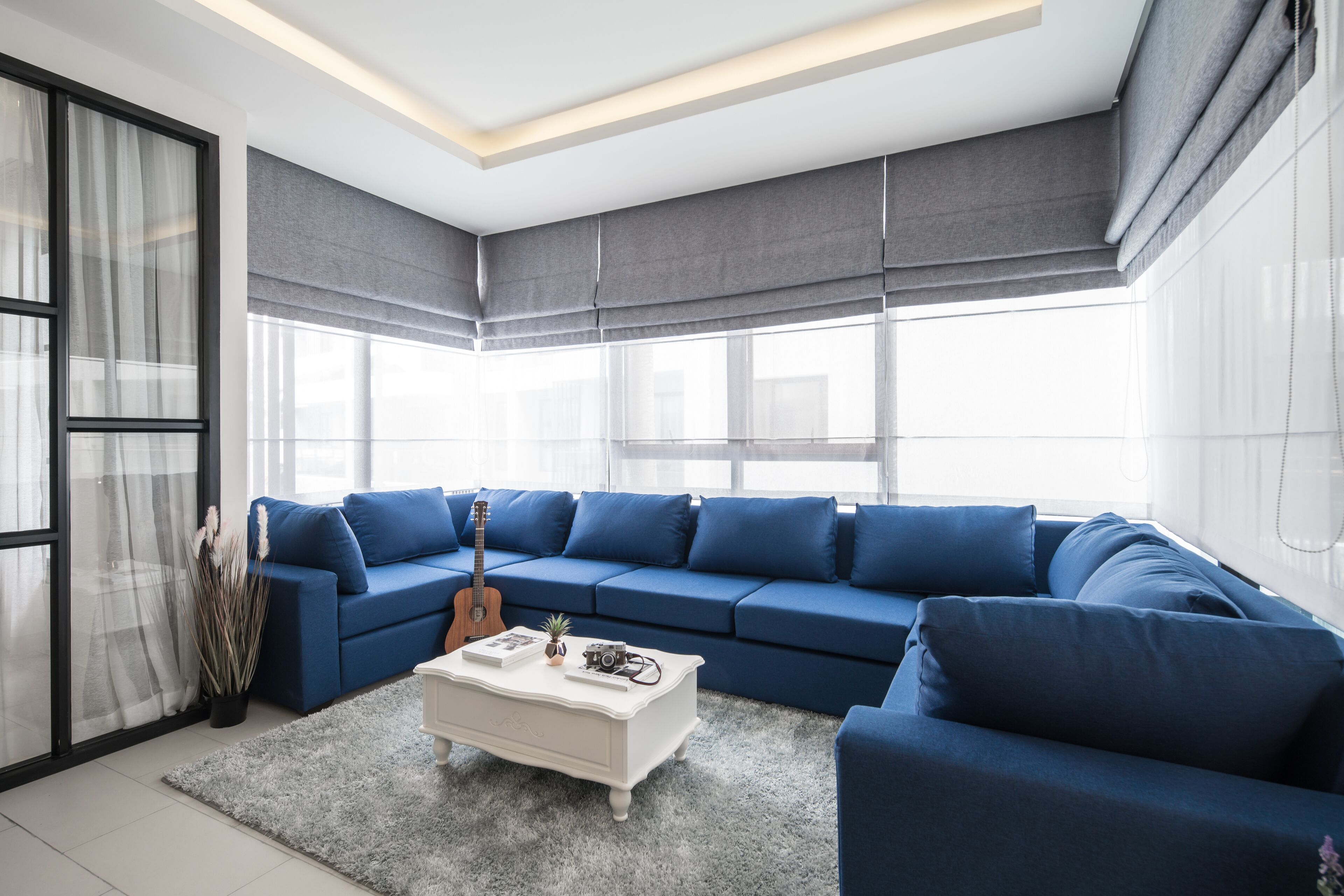A modern living room with a blue sectional sofa, a white coffee table, and a large window with sheer curtains.