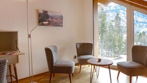 Ein modernes Wohnzimmer mit Holzboden, einer stehenden Stehlampe, zwei gemusterten Sesseln, einem kleinen runden Tisch und einem großen Fenster mit Bergblick.