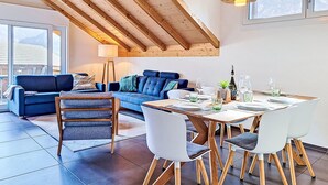 Ein modernes Wohnzimmer mit blauem Sofa, einem Holzespeisetisch und Stühlen, einer Lampe und Bergblick durch große Fenster.
