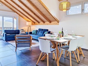 Un salon moderne avec un canapé bleu, une table à manger en bois et des chaises, une lampe, et une vue sur les montagnes à travers de grandes fenêtres.