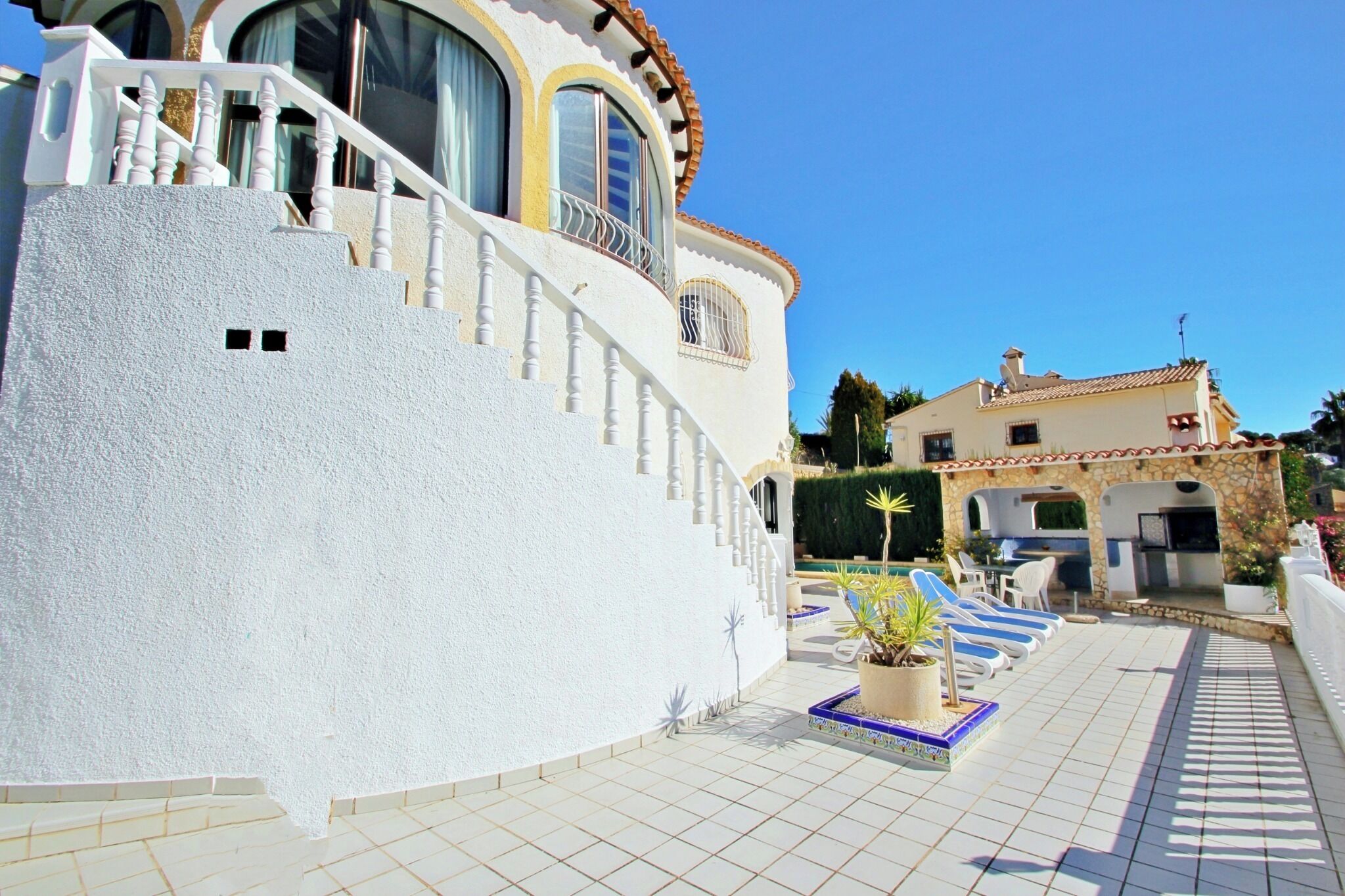 A white building with a curved facade, a balcony with a railing, and a tiled patio area with lounge chairs and a small pool.