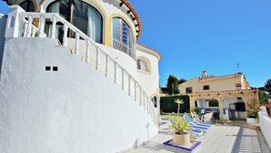 En hvid bygning med en buet facade, en balkon med gelænder og en fliseterrasse med liggestole og en lille pool.
