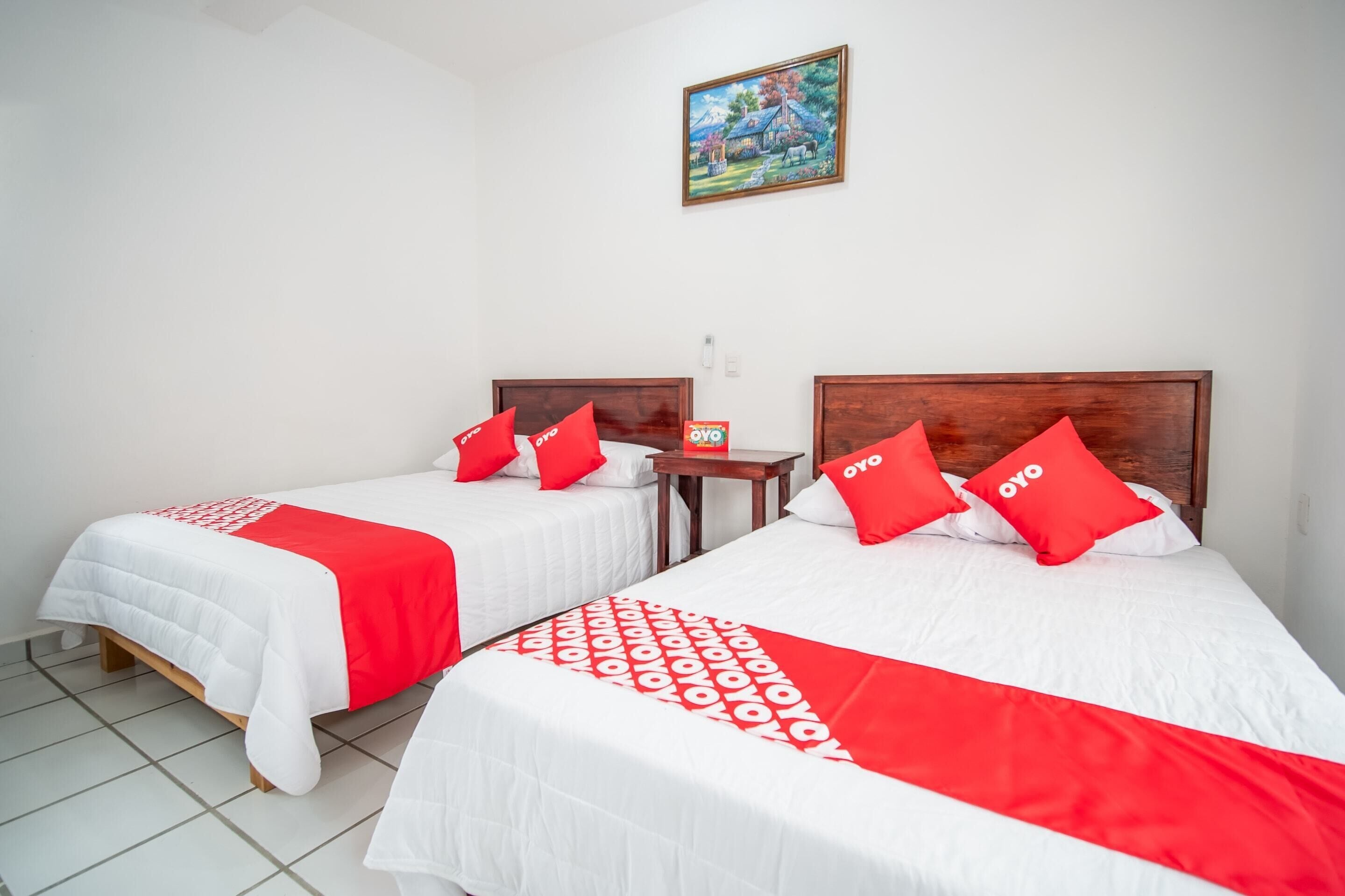 A hotel room with two beds, each with a wooden headboard, white bedding, and red accents. There is a small table between the beds, and a painting hangs on the wall above.