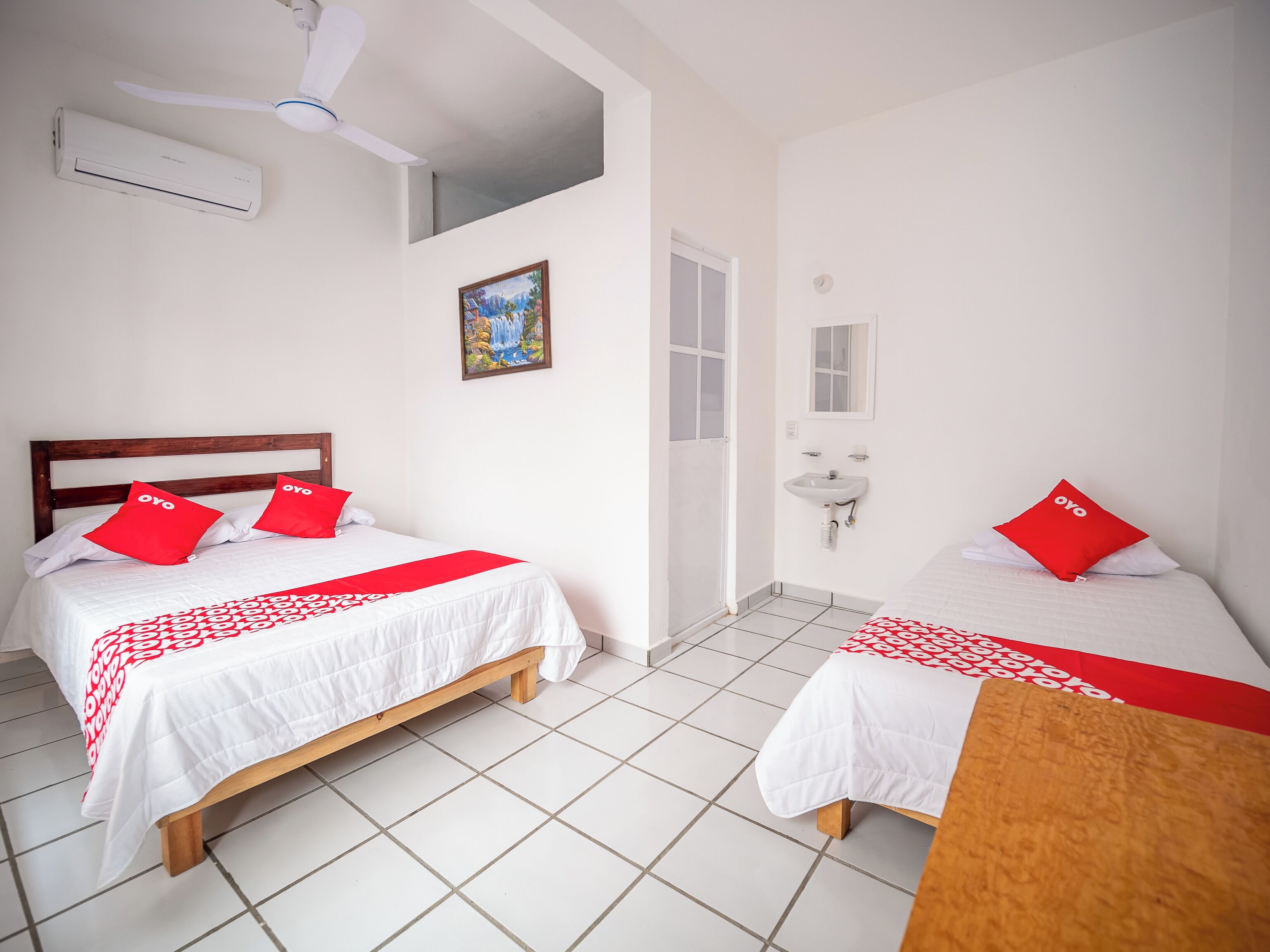 Una camera d'albergo con due letti, un ventilatore a soffitto, un condizionatore d'aria, un quadro, un lavandino e uno specchio.