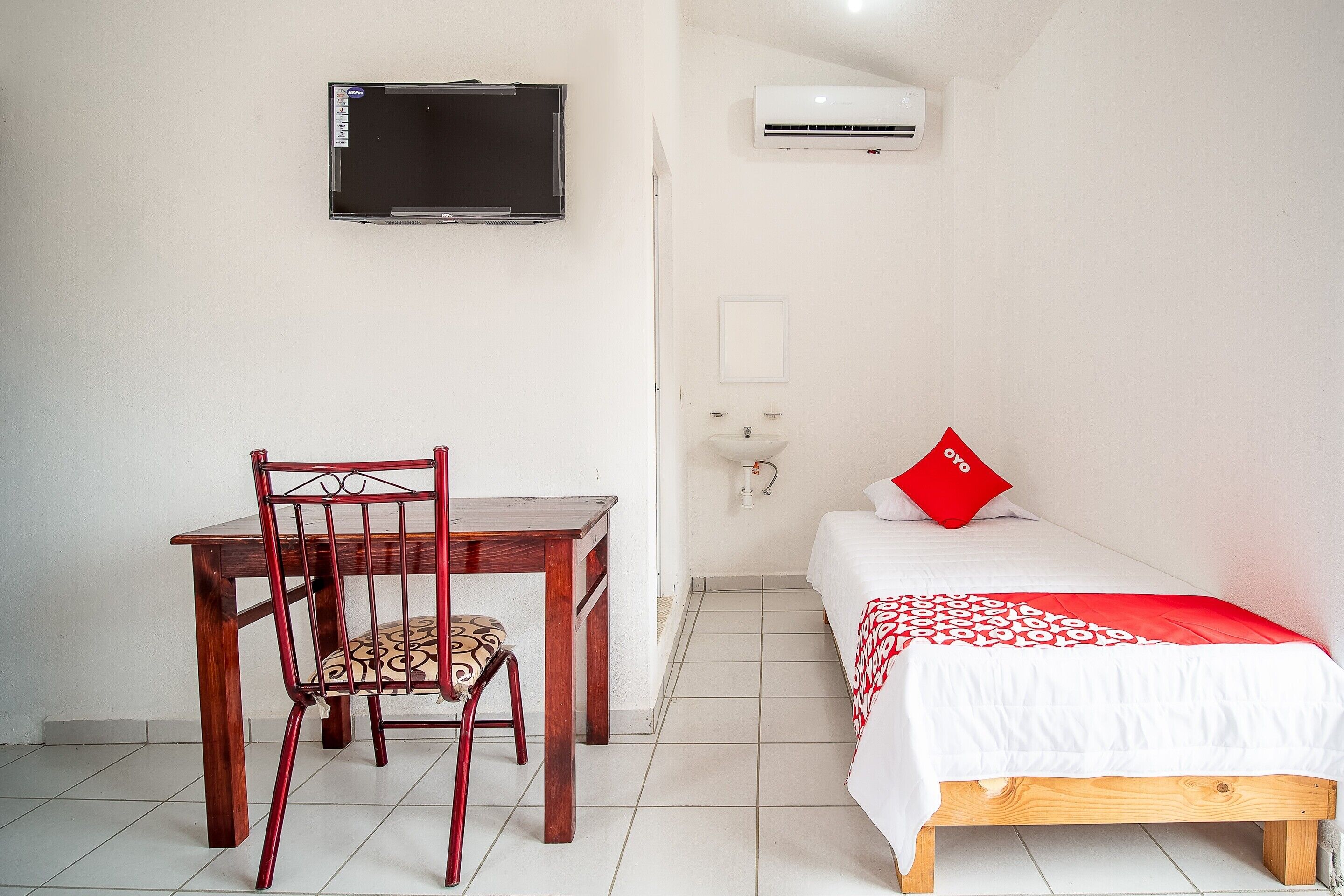 Una habitación pequeña y limpia con un escritorio de madera, una silla, una cama con almohada roja, un televisor y aire acondicionado.