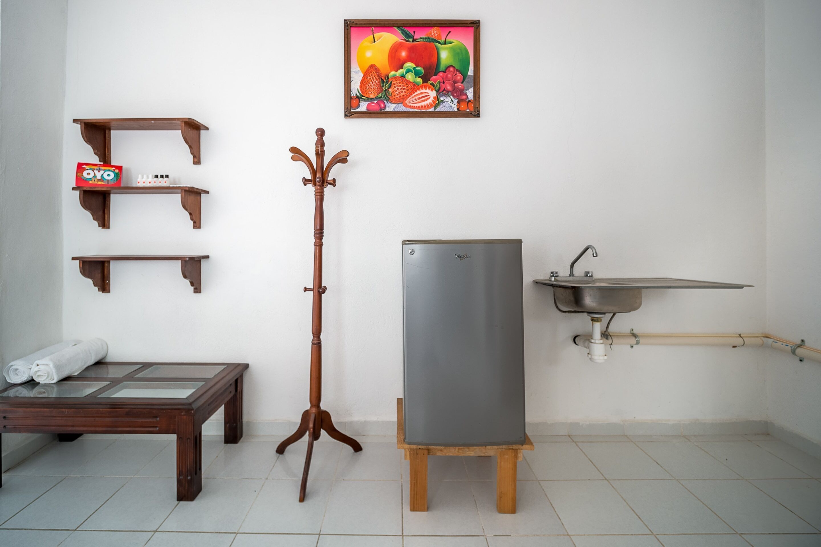 A room with a wooden table, a rolled-up towel, a stand with a box, a framed painting, a tall wooden stand, a small wooden stool, and a sink with pipes.