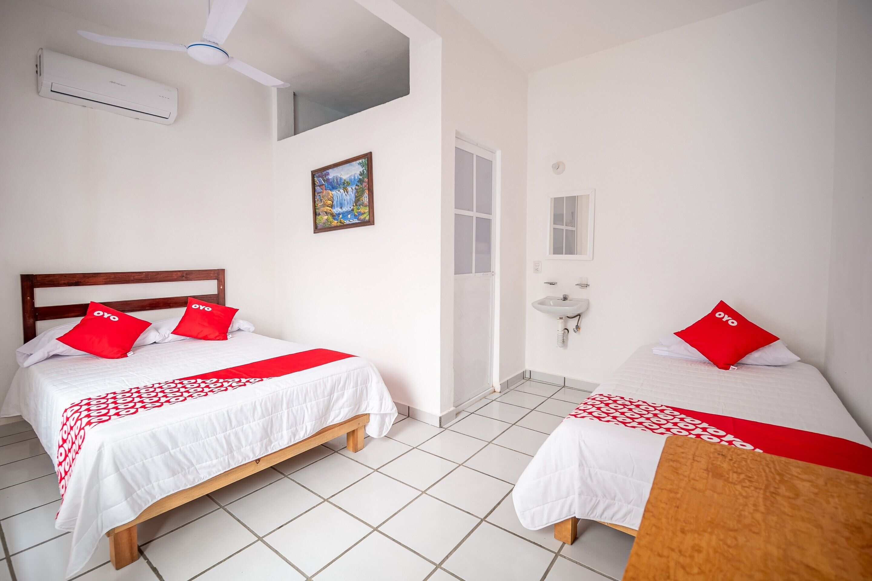 Une chambre d’hôtel avec deux lits, chacun recouvert d’une couvre-lit rouge et blanche assortie aux coussins. On y trouve également un ventilateur de plafond, un appareil de climatisation et un tableau accroché au mur.