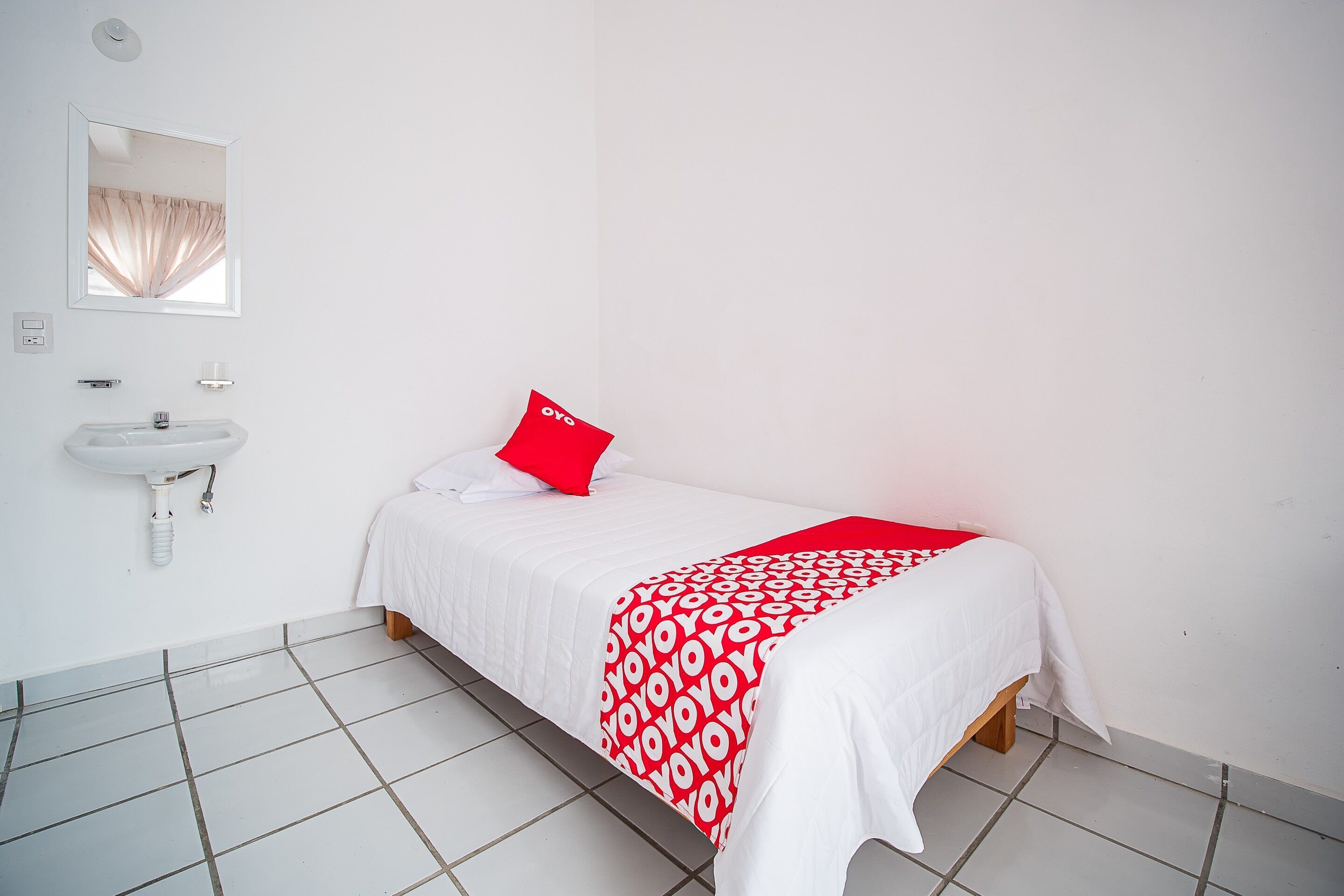 Una camera da letto con un letto, un cuscino rosso, una coperta bianca e un lavandino bianco, oltre a uno specchio appeso al muro.