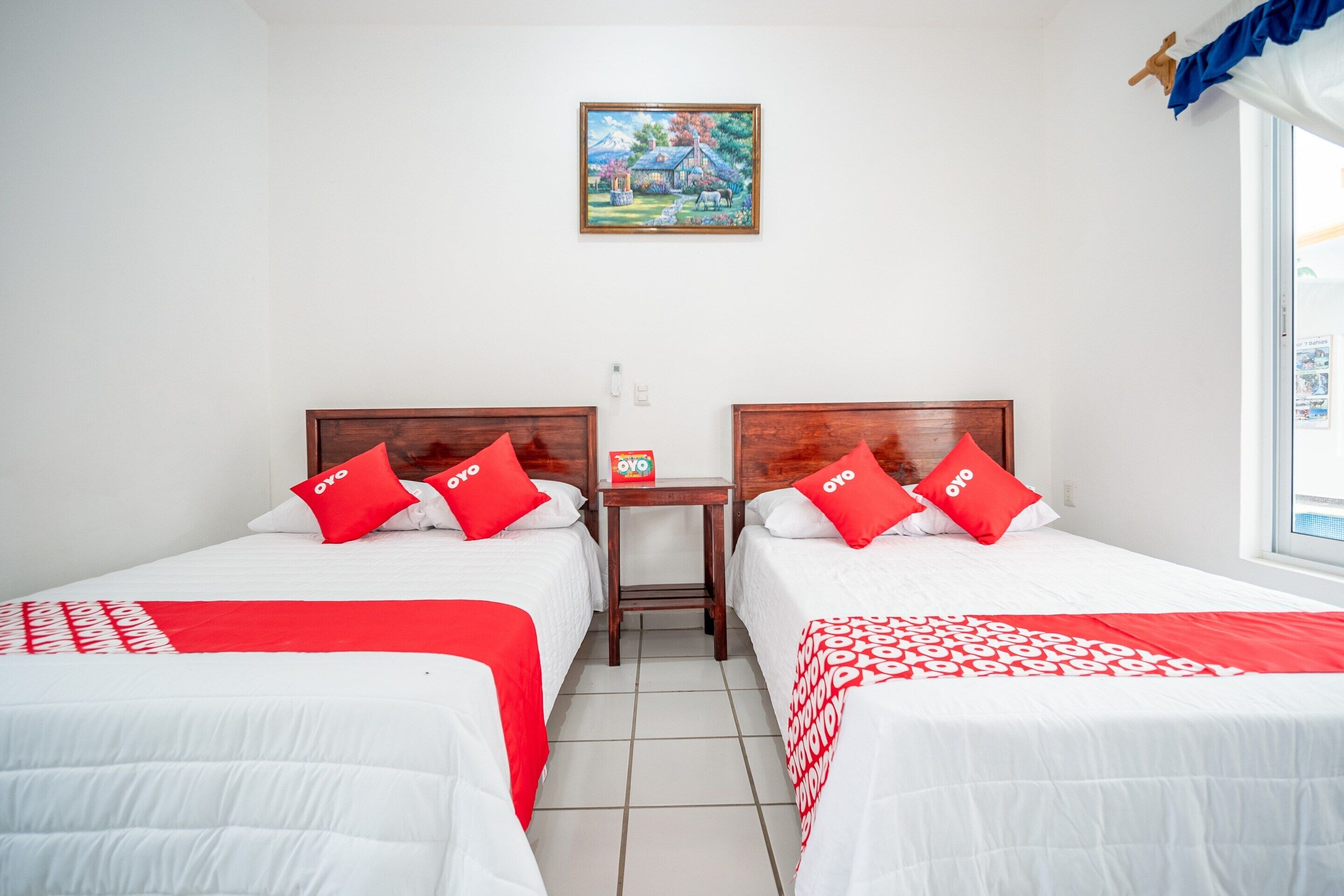 Quarto de hotel com duas camas, cada uma com roupa de cama vermelha e branca, uma pequena mesa com um objeto vermelho e um quadro na parede.