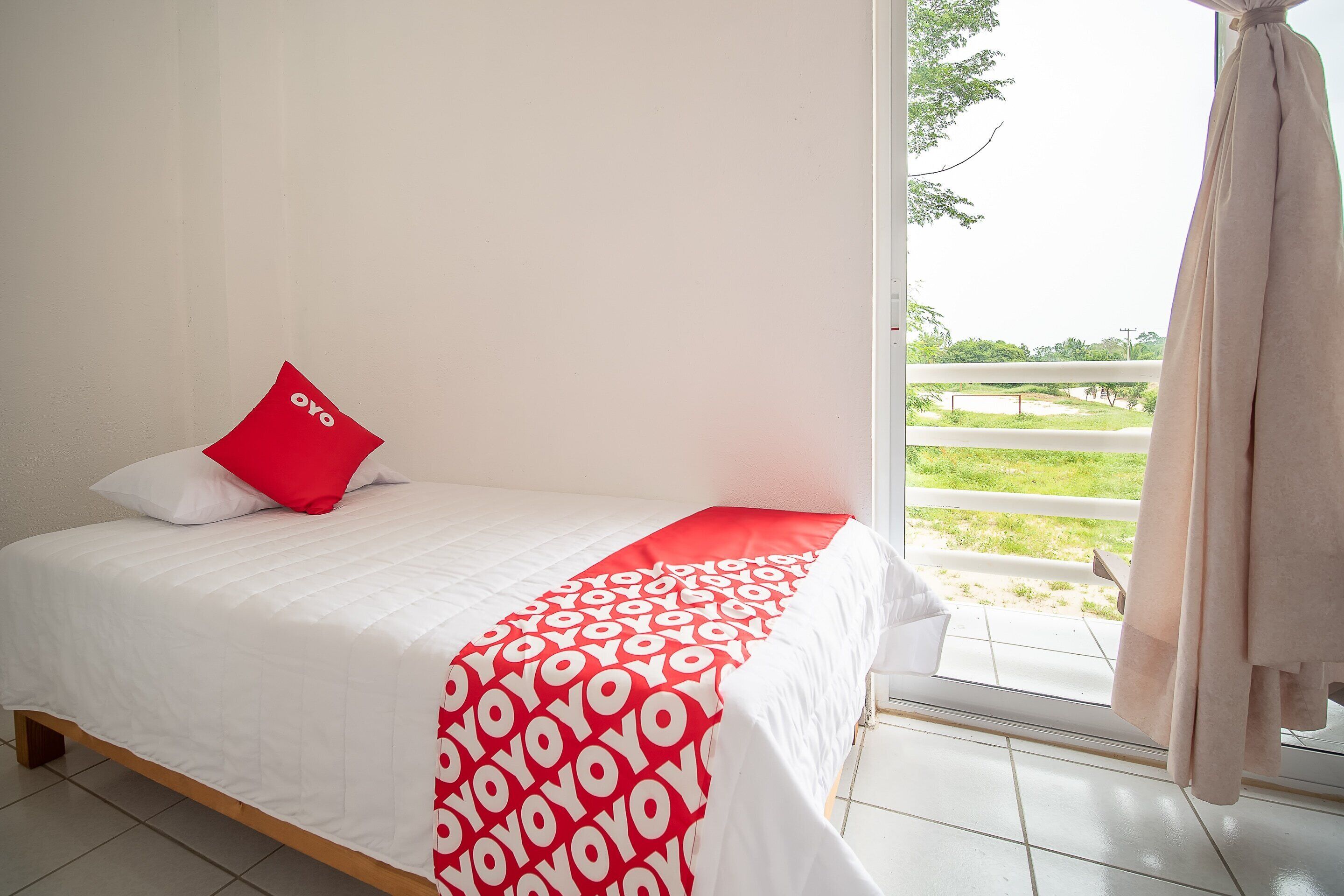 A bed with a white and red bedspread, a red pillow, and a view of a green landscape through a window.