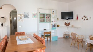 Un coin repas avec une table en bois et des chaises à motifs orange, une cuisine avec des meubles turquoise et un téléviseur posé sur une étagère, et un fauteuil en osier recouvert d’une nappe blanche.