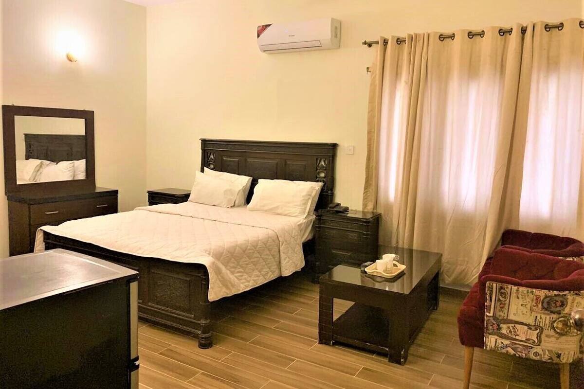 A hotel room with a bed, bedside tables, a chair, a desk, and an air conditioning unit.