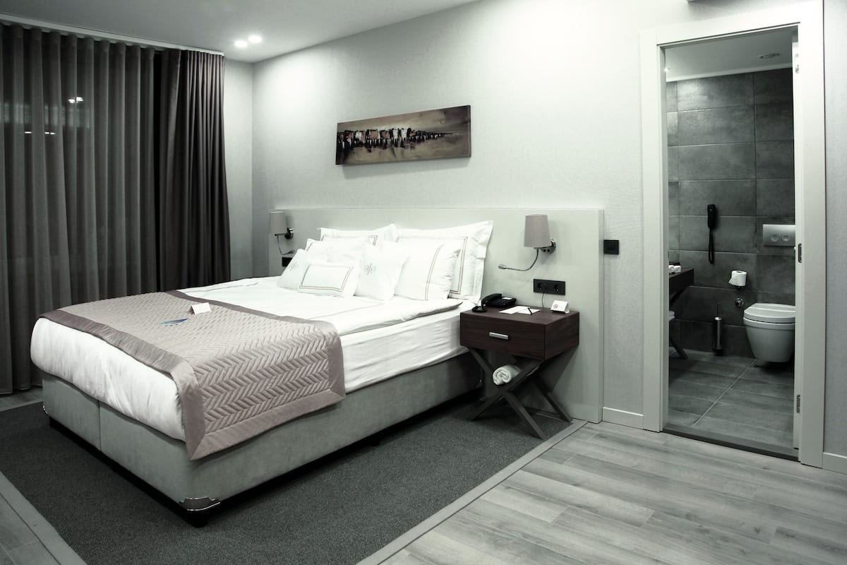 A modern hotel room with a large bed, a bedside table, a nightstand, a wall-mounted telephone, and a view into the bathroom.
