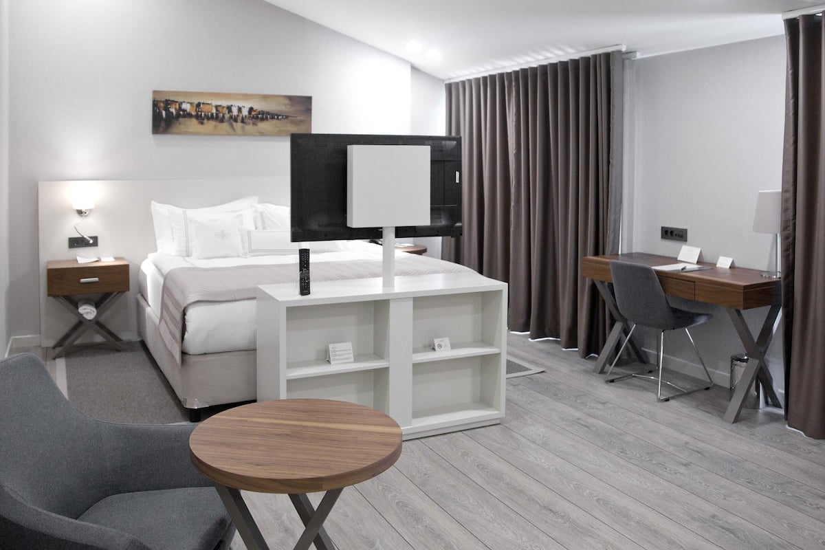 A modern hotel room with a bed, a desk, a chair, a small table, and a TV.