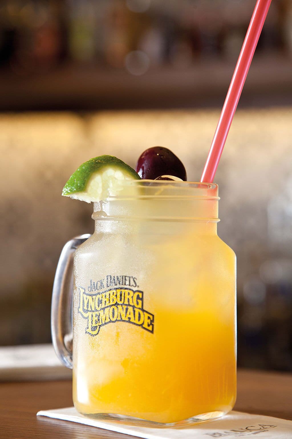 A mason jar with a red straw, containing a yellow beverage, garnished with a lime slice and a cherry, labeled "Jack Daniel's Ingeburg Lemonade."