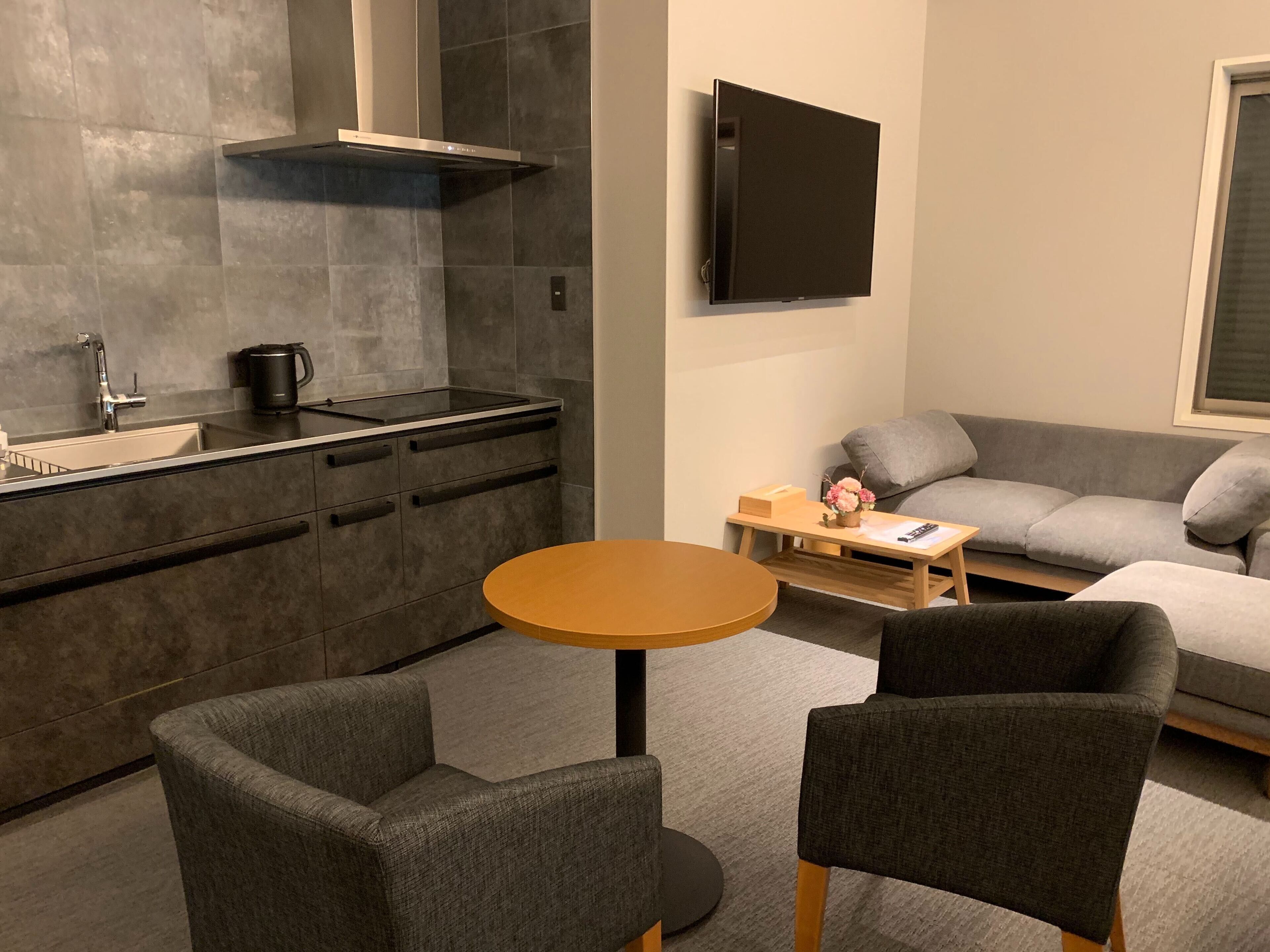 A modern kitchen with a built-in stove, a coffee maker, and a sink. There is a round wooden table with two chairs in front of a grey sofa. A flat-screen TV is mounted on the wall above the sofa.