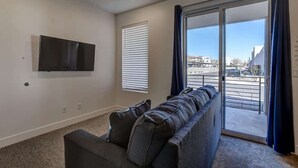 A living room with a sofa, a flat-screen TV mounted on the wall, and a sliding glass door leading to an outdoor area. - The Lofts #36 | Townhome Pool Resort (Saint George)