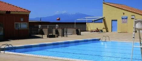 Ein Pool mit freiem Blick auf Berge im Hintergrund, einem überdachten Sitzbereich und einem Gebäude mit rotem Dach.