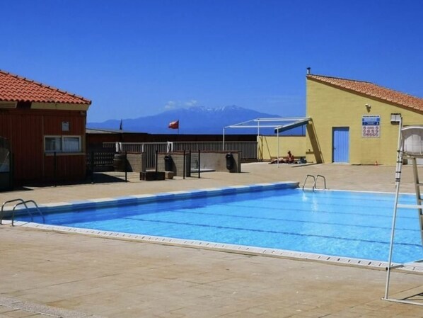 Une piscine offrant une vue dégagée sur les montagnes en arrière-plan, un espace de repos couvert et un bâtiment avec un toit rouge.
