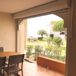 An indoor covered terrace with a wooden table and chairs, a view of a landscaped garden with trees and a building in the background. - Ideal family, 3 rooms ground floor, terrace, in peninsula Coudalère (Le Barcarès)