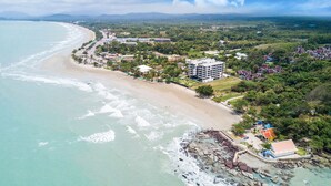 An aerial view of a coastal area with a beach, a large hotel with multiple floors, and surrounding greenery. - Gorgeous Sunset View ❤︎ Mae Phim Beachfront Condo (Kram)