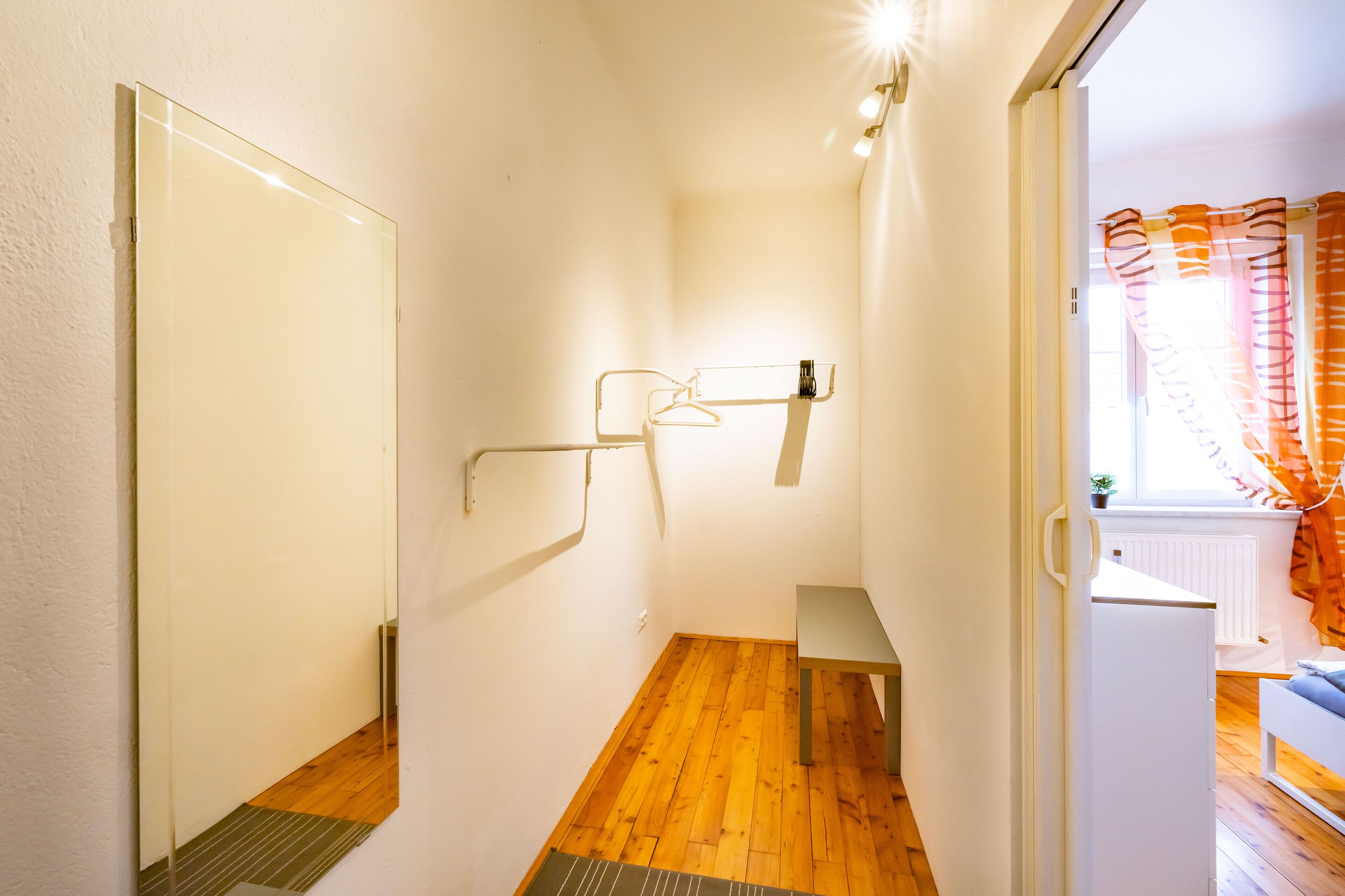 A hallway with a wooden floor, a mirror, a coat rack, and a door leading to a room with a window and curtains.