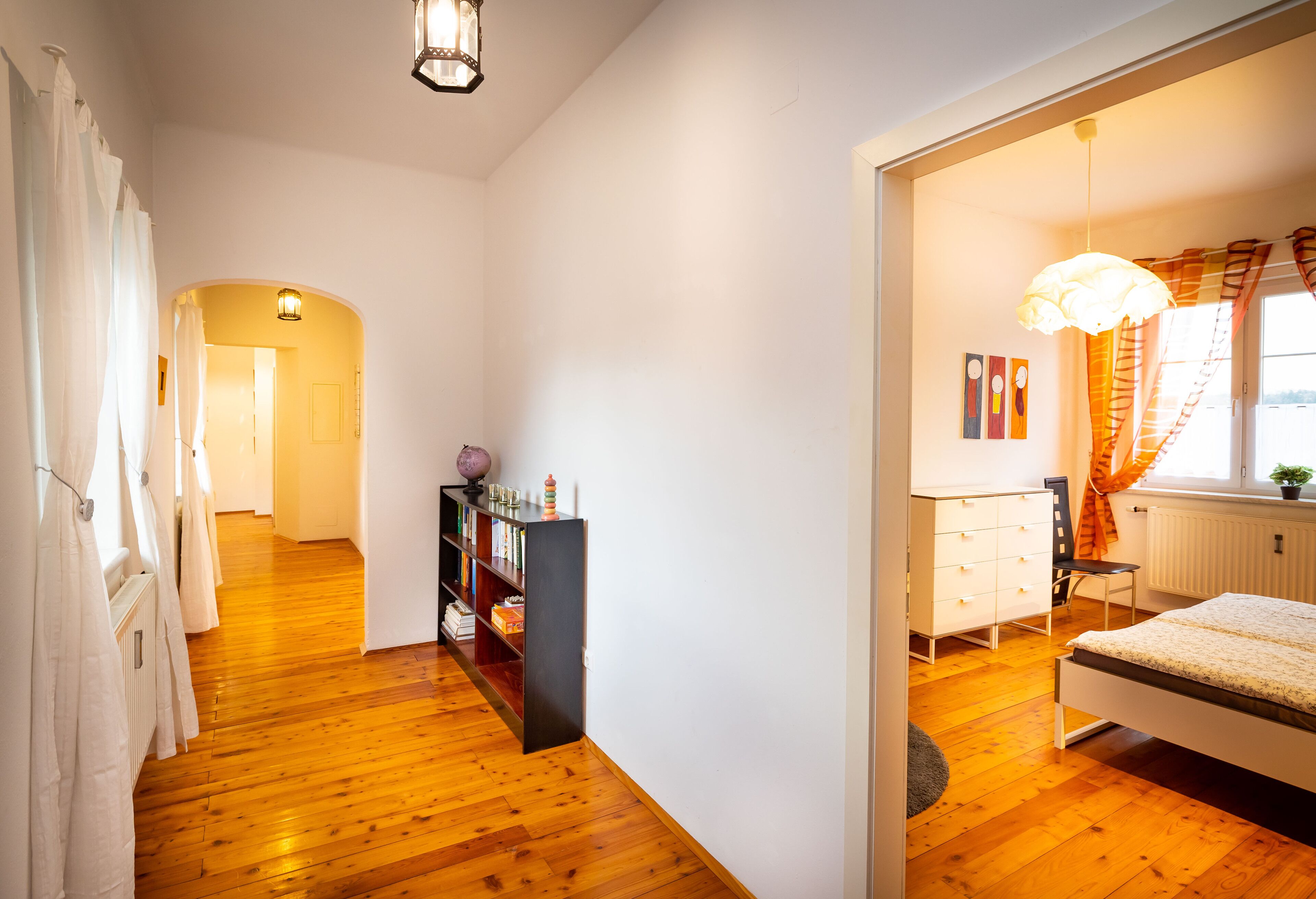 Un dormitorio con una cama, un aparador y un escritorio con una silla. Se ve un pasillo que conduce a otra habitación.