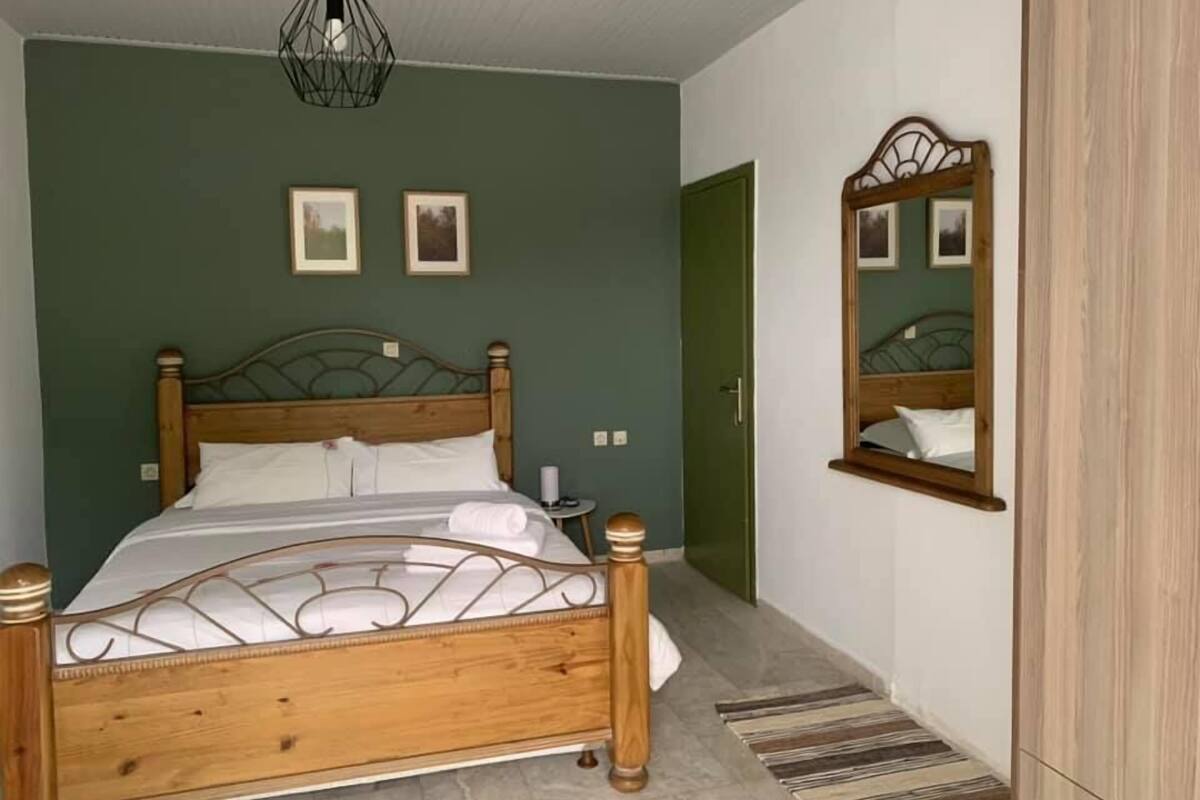 A bedroom with a wooden bed, a green wall, two framed pictures, a mirror, and a small bedside table.